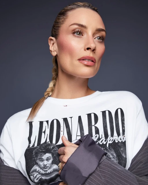 Portrait of a blonde adult female wearing a Leonardo DiCaprio graphic t-shirt in a photo studio with a dark grey background.