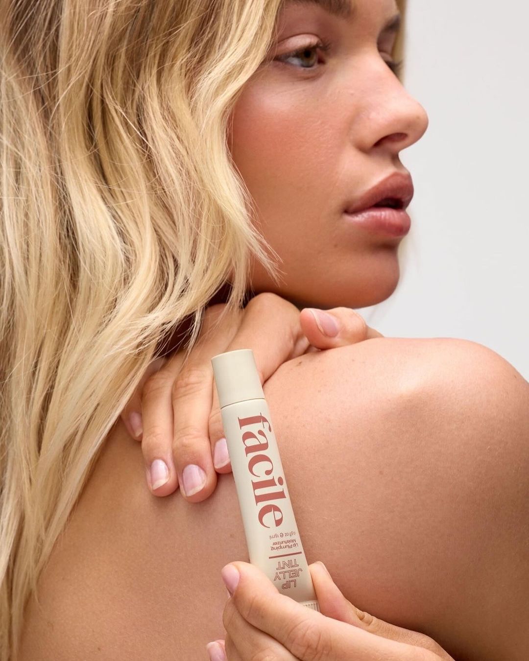 Close-up shot of an adult woman with fair skin and blonde hair holding a tube of 'facile' lip jelly tint against a white background.