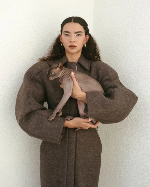 A young woman with curly hair, wearing a brown wool coat, holds a grey hairless cat in her arms against a light grey wall background.