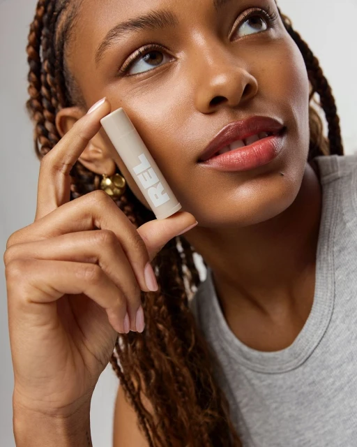 Woman holding a cosmetic stick near her face. The product features 'REL' printed on the side. The model has braided hair and wears a grey top.