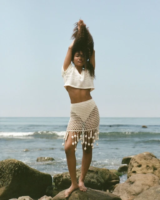 A woman with curly hair stands barefoot on rocks by the ocean, wearing a knitted top and skirt against a clear blue sky.
