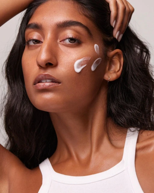 Close-up of a young adult woman with dark hair and skin wearing a white tank top and posing with face cream on her cheek.
