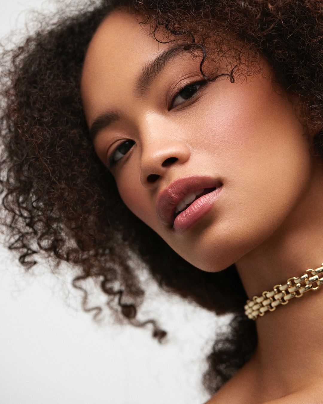Close-up portrait of a young adult woman with curly dark hair, wearing a gold choker against a soft white background. Beauty shot, angled view.