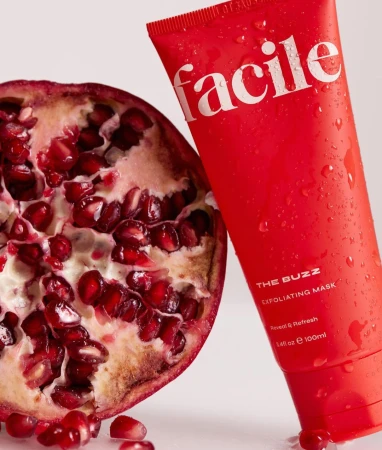 A red exfoliating mask tube next to a halved fresh pomegranate with glistening arils against a white background