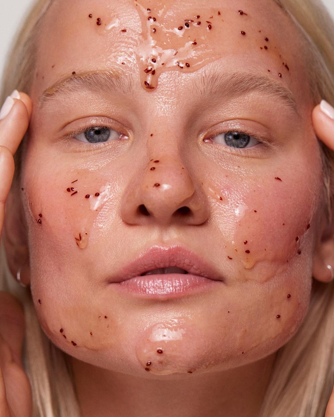 Close-up of a young woman's face covered in a clear, gel-like mask with small brown particles; applying mask with fingertips in studio lighting.