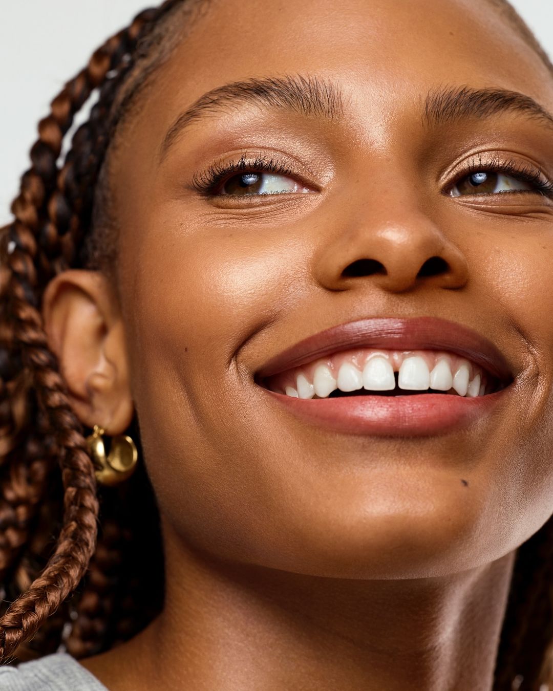 Close-up of a smiling African-American woman with brown skin and braided hair, accessorized with gold hoop earrings, set against a neutral backdrop.
