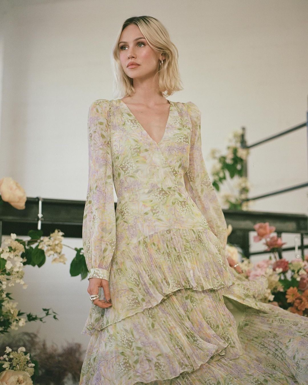 A woman in a flowing floral dress stands surrounded by flowers in a bright indoor space with filtered light, looking away from the camera.