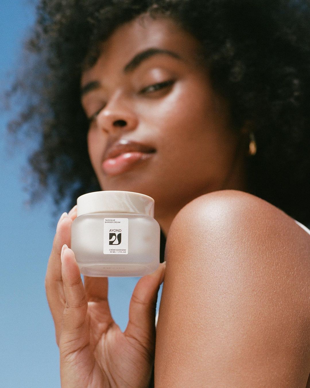 A young adult Black woman holds a jar of skincare product against a blue sky, showcasing a beauty and wellness aesthetic with natural lighting.
