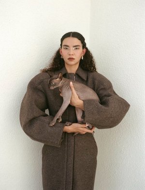 Adult woman in a brown coat holds a hairless cat in front of a white wall, wearing hoop earrings, and with long curly hair.