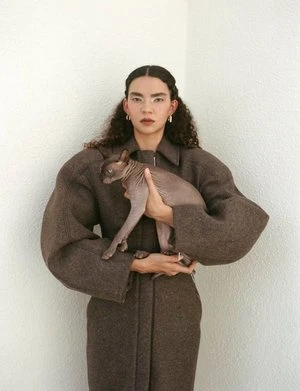 Adult woman in a brown coat holds a hairless cat in front of a white wall, wearing hoop earrings, and with long curly hair.