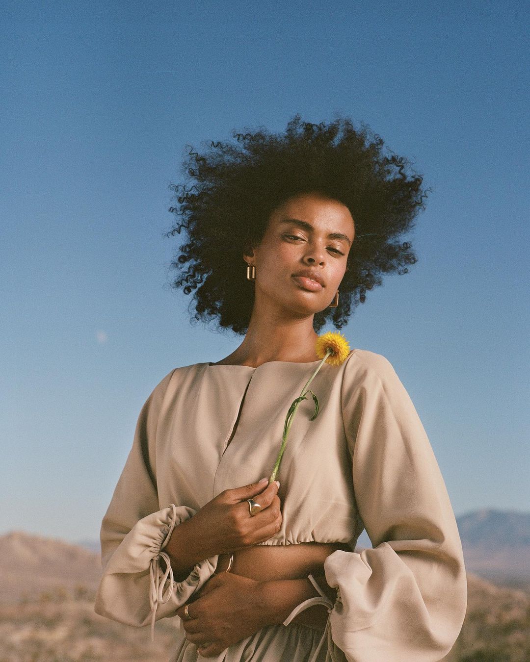 Portrait of a woman with afro holding a yellow flower in a desert landscape with a clear blue sky. She is wearing a beige two-piece outfit.