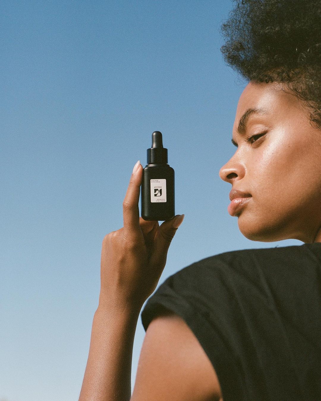 Woman in black holds cosmetic dropper bottle against a clear blue sky in a beauty or skincare product shoot.