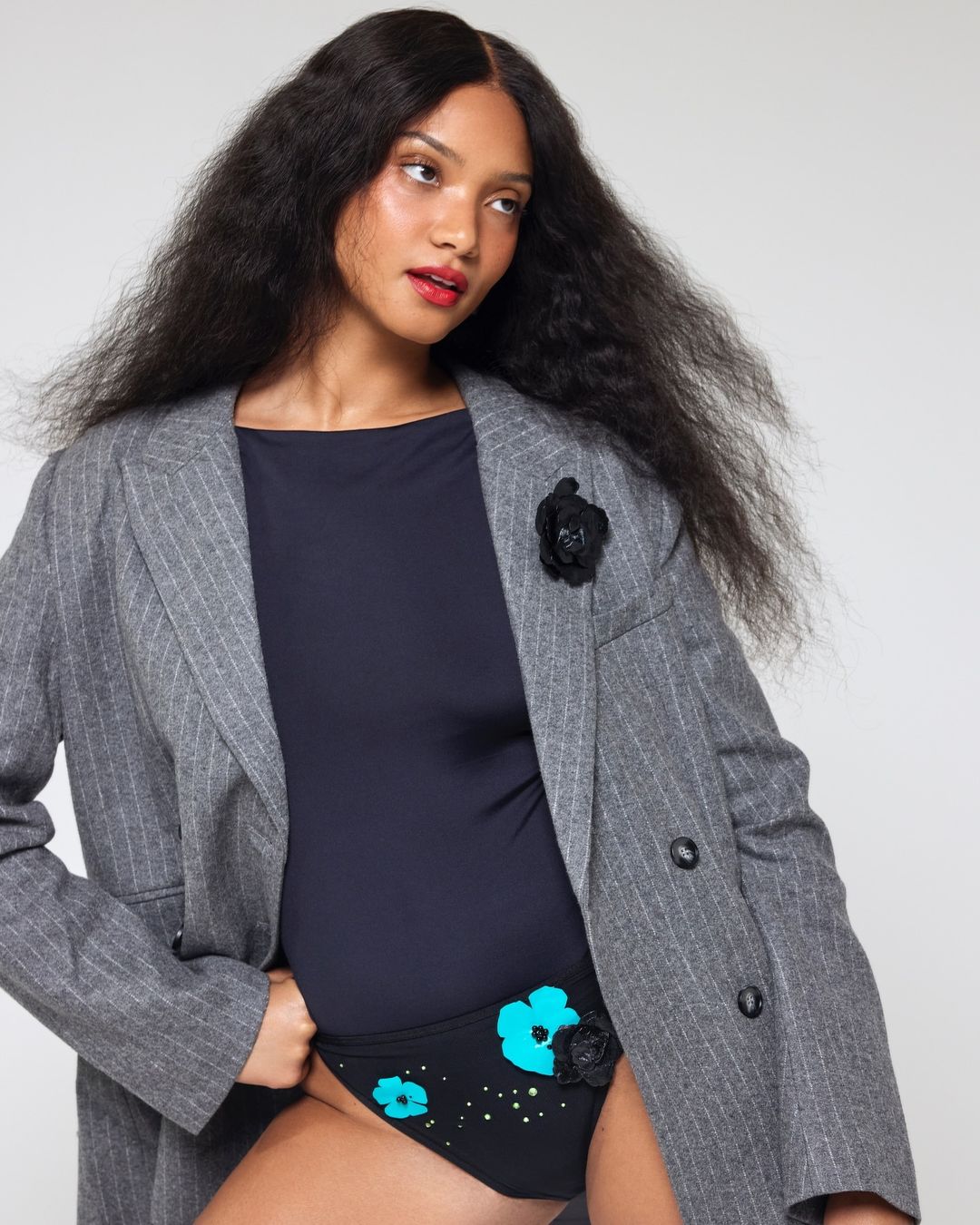 Woman wearing a striped blazer and swimsuit bottom with flower details in a studio setting with a soft, neutral-toned backdrop.