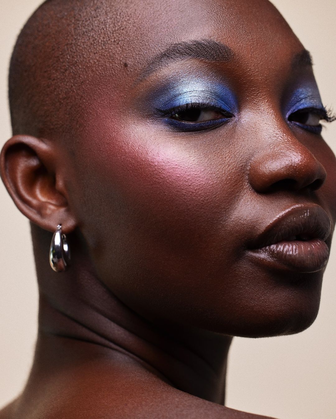 Close-up portrait of a Black woman with shimmering blue eyeshadow and pink blush, wearing small silver hoop earrings.