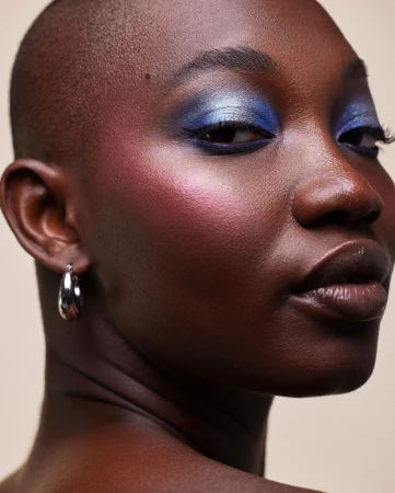 Close-up portrait of a Black woman with shimmering blue eyeshadow and pink blush, wearing small silver hoop earrings.