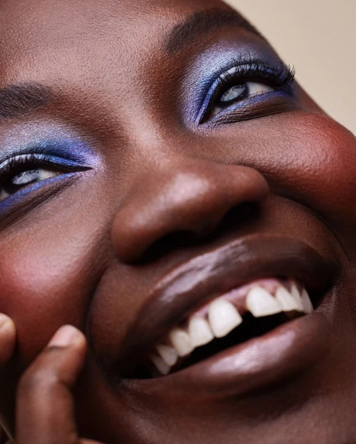 Close-up portrait of a smiling Black woman with iridescent blue eyeshadow, glossy lips, and dark skin against a neutral background.