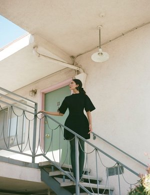 Adult female in a short black dress standing on metal stairs outside of a building with a pale-green door and white walls in daylight.