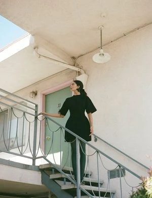 Adult female in a short black dress standing on metal stairs outside of a building with a pale-green door and white walls in daylight.