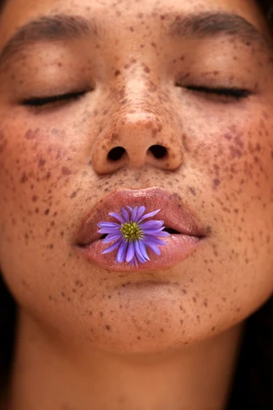 Close-up of a woman's face with freckles and closed eyes, holding a purple flower between her lips in an indoor setting with soft lighting.