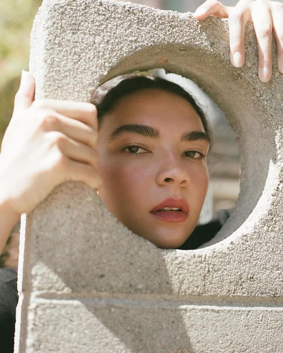 Close-up of a mixed-race female adult looking through a hole in a concrete block, with neutral expression and natural light.