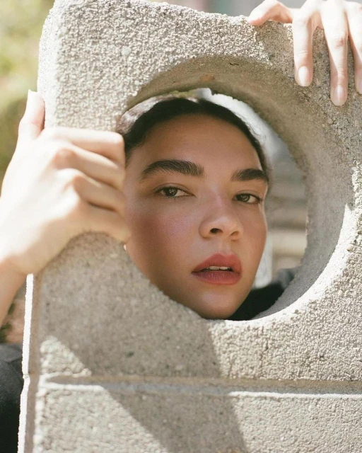 Close-up of a mixed-race female adult looking through a hole in a concrete block, with neutral expression and natural light.