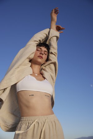 Young woman poses outdoors with arms raised against a clear blue sky wearing a white crop top and beige linen pants and jacket.