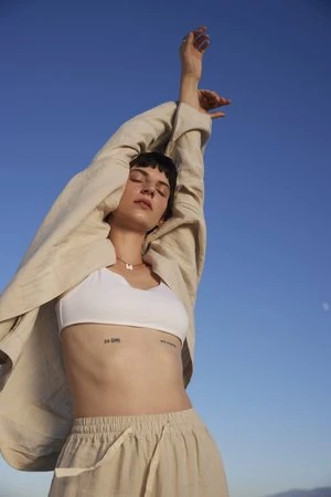 Young woman poses outdoors with arms raised against a clear blue sky wearing a white crop top and beige linen pants and jacket.