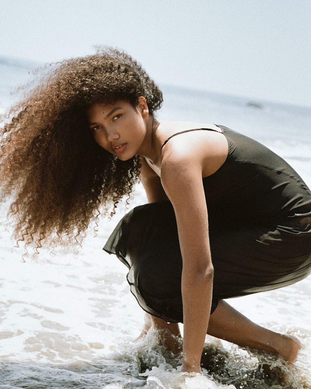 Woman with voluminous curly hair wearing a dark slip dress squatting at the water edge on a sandy beach