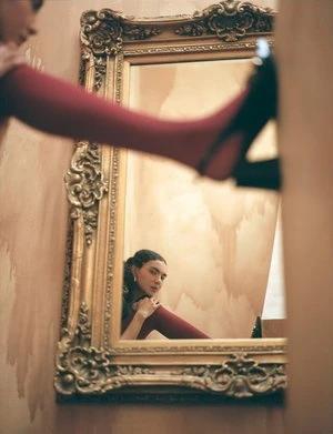 Woman reflected in a gold ornate mirror wearing red tights, framed by the legs of another person in a high heel shoe.