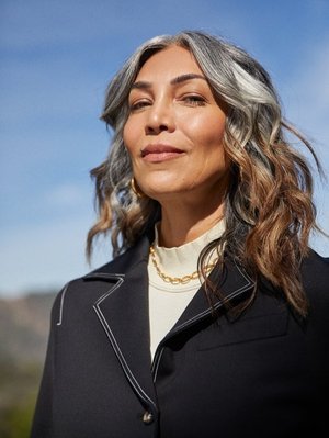 Portrait of a middle-aged woman with graying hair wearing a black jacket and gold necklace, set against a bright blue sky.
