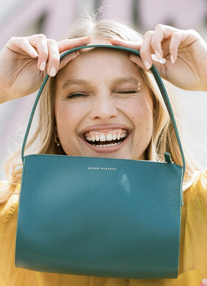 Close-up portrait of a smiling blonde woman holding a teal handbag up to her face with both hands against an out-of-focus background.