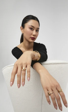 Portrait of a woman wearing gold jewelry and rings resting her arms on a white chair against a plain white background