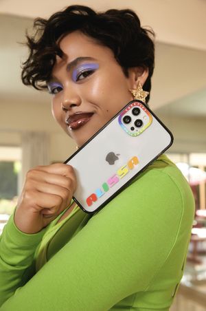 Smiling woman with short hair holds up a white phone with colorful custom lettering, wearing bright green top indoors.