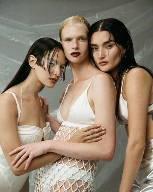 Three women in white outfits pose together in front of a blurry, light grey backdrop. Fashion editorial photography.