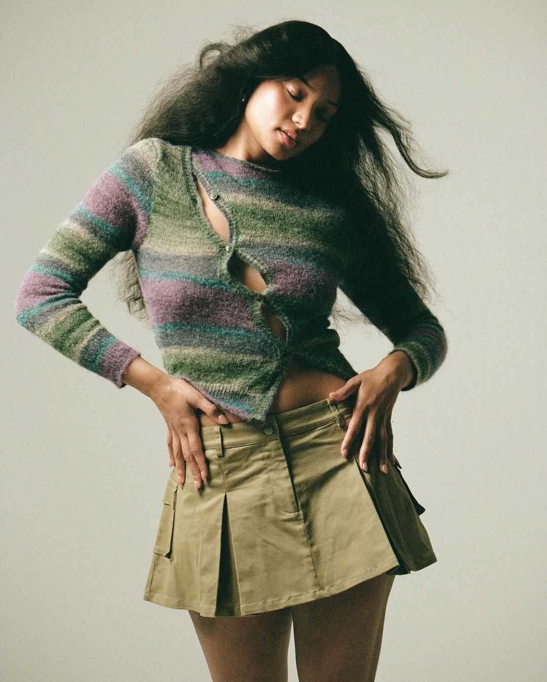 A fashionable young woman poses indoors, wearing a horizontally striped sweater with a khaki mini skirt, long dark hair, against a neutral backdrop.