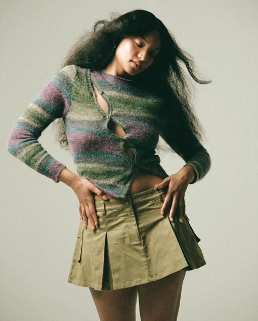 A fashionable young woman poses indoors, wearing a horizontally striped sweater with a khaki mini skirt, long dark hair, against a neutral backdrop.