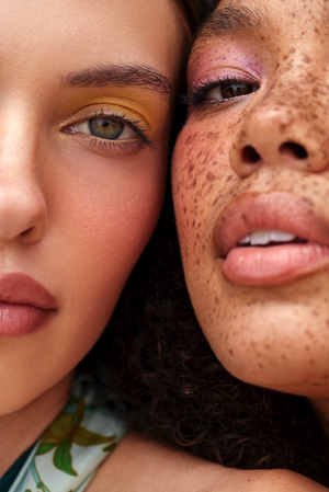 Close-up studio shot of two diverse women showcasing colorful eyeshadow makeup. One woman has freckles, and both have pink lips.