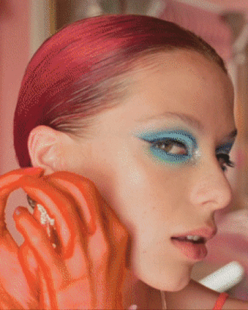Close-up portrait of a woman with slicked-back red hair, vibrant blue winged eyeshadow, and bright orange gloves touching her ear.