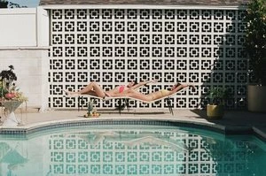 Two women in swimsuits lounge by a pool with patterned privacy screen and potted plants, capturing a summer leisure scene.