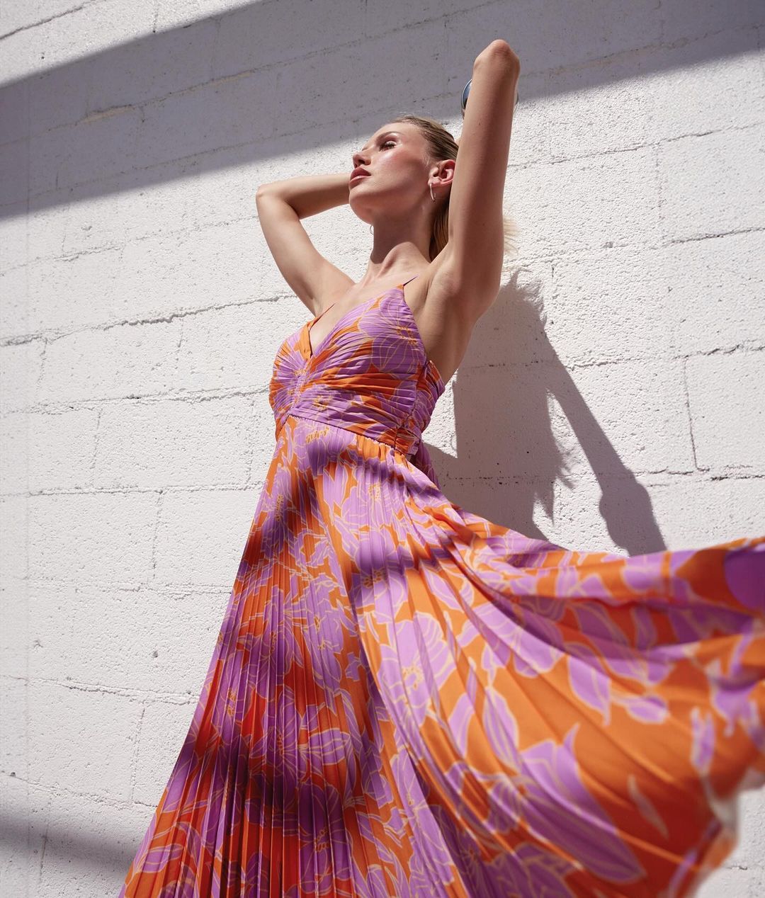 Woman in flowing orange and purple floral gown poses against a textured white wall bathed in sunlight, casting shadows and highlights.