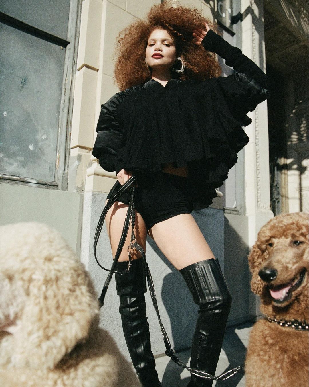 Adult woman with curly hair in a black dress and high boots poses with two standard poodles on a city street with buildings in the background.