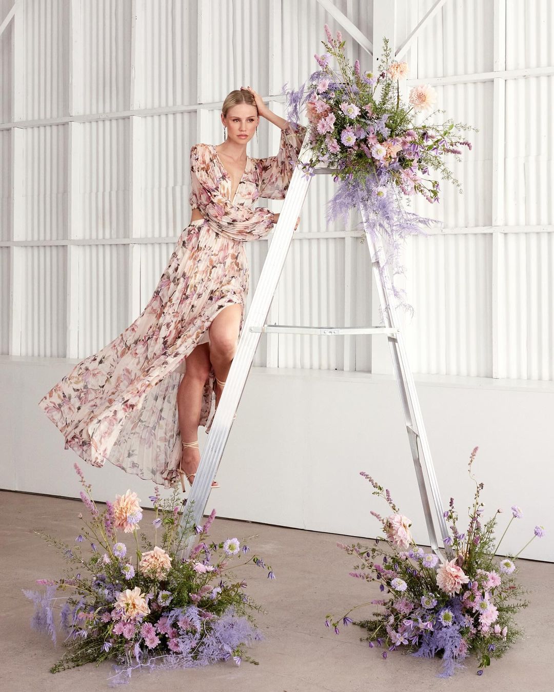 A blonde woman in a long floral dress poses on a silver ladder surrounded by floral arrangements, bathed in soft, diffused light.