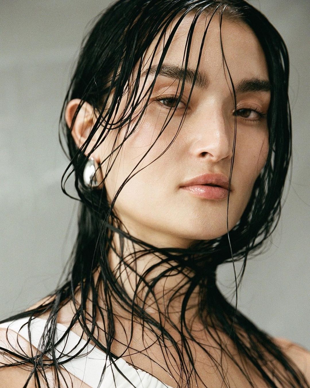 Close-up portrait of a woman with wet dark hair clinging to her face, wearing a white top and silver hoop earrings.