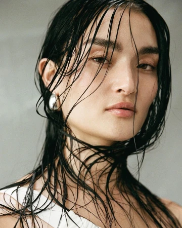 Close-up portrait of a woman with wet dark hair clinging to her face, wearing a white top and silver hoop earrings.
