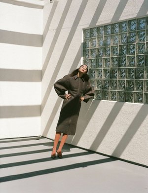 A stylish woman poses indoors near a glass block window, casting geometric shadows across her and the stark white walls.