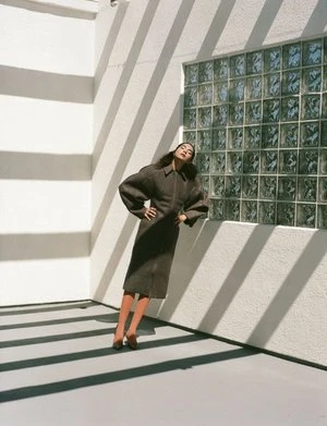 A stylish woman poses indoors near a glass block window, casting geometric shadows across her and the stark white walls.