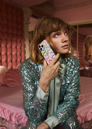 Person holding a colorful heart-patterned iPhone case while posing in a room with a pink bed, tufted headboard, and a sequined jacket.