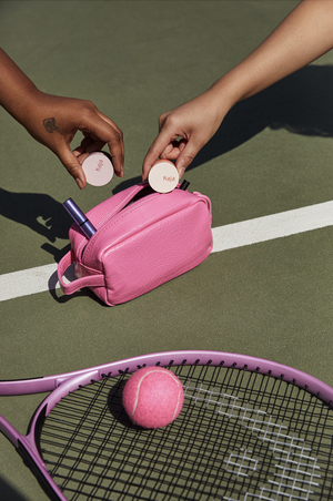 Two hands reach for makeup compacts above a pink cosmetics bag with tennis racket and ball on a green tennis court.