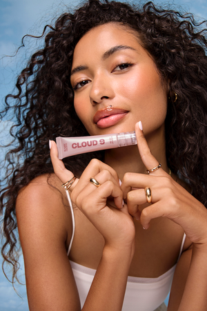 Close-up portrait of a woman holding a lip gloss tube labeled Cloud 9 against a light blue sky background.