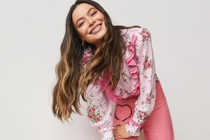 Portrait of Miranda Cosgrove smiling and wearing a floral top and pink skirt against a neutral background in an eye-level medium shot.